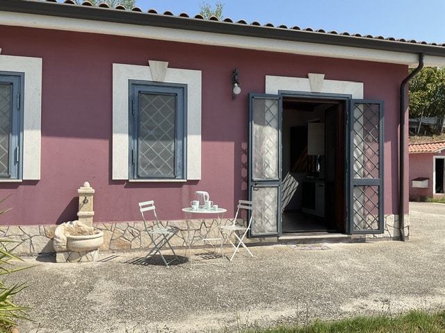 A quaint outdoor seating area is depicted, featuring a small round table with two chairs. The entrance to the dwelling is framed by double doors, while a decorative fountain adds a charming touch. The building's exterior is painted in a warm shade of pink.