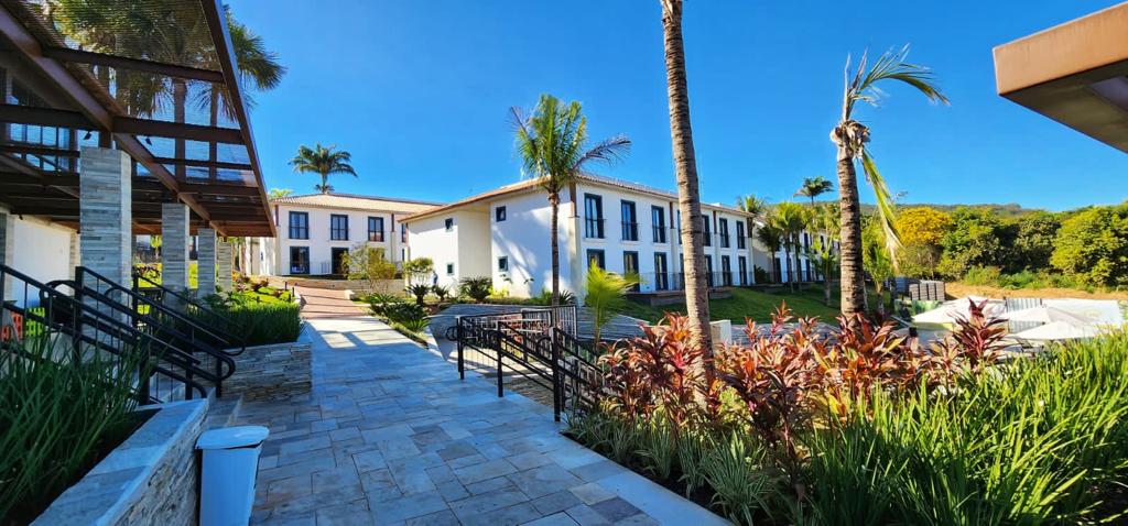 The image depicts a serene outdoor area with well-maintained gardens featuring tropical plants and palm trees. A stone pathway leads to a white multi-story building, with several large windows visible. Bright blue skies complement the inviting atmosphere.