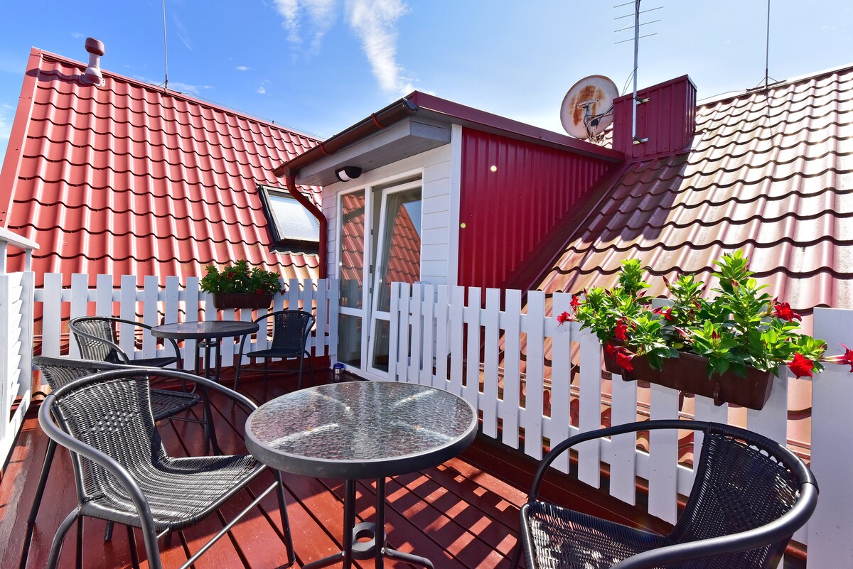 An outdoor balcony is presented with two black wicker chairs and a round glass table. Flower boxes with vibrant blooms adorn the white picket fence. The red-tiled rooftops provide a charming backdrop under a clear blue sky.
