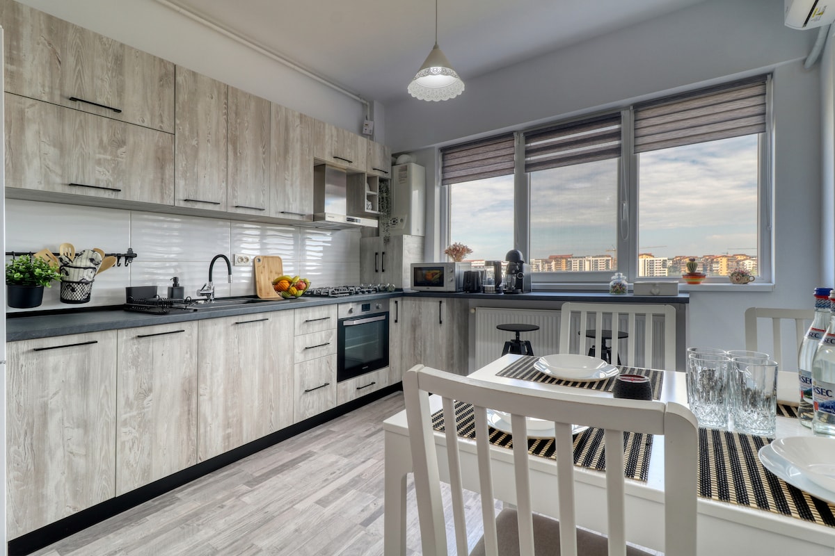 The kitchen area features modern cabinetry with a neutral finish, complemented by a black countertop. Appliances include a gas stove, oven, and microwave. Large windows provide natural light and views of the surroundings. A dining table with two chairs is set for a meal.