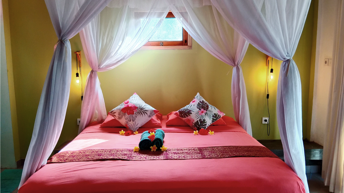A spacious bed is draped with a sheer canopy, featuring vibrant floral-patterned pillows. Soft lighting is provided by wall sconces on either side. A refreshing atmosphere is created by the light streaming through the window, complementing the warm tones of the walls.