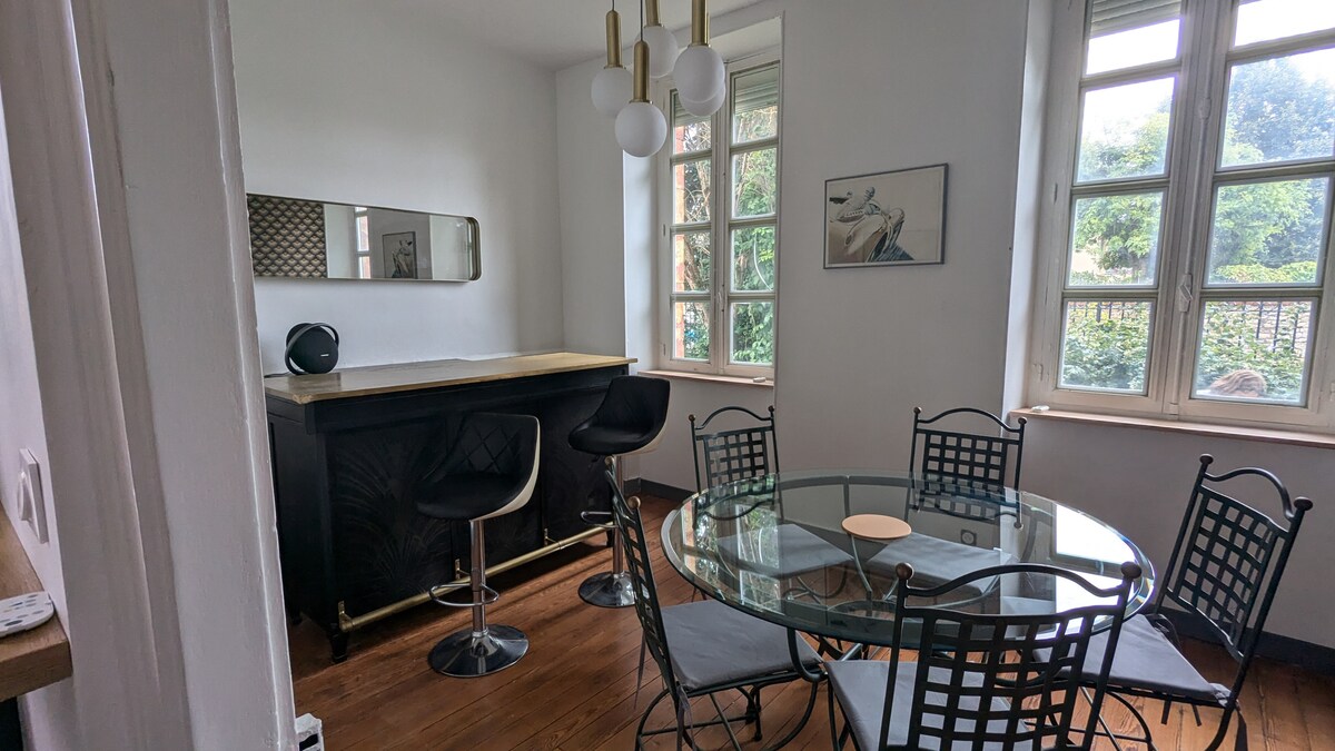 A dining area features a round glass table surrounded by black wrought iron chairs. A sleek bar counter is present against one wall, with modern pendant lights hanging above. Natural light enters through large windows, illuminating the space.