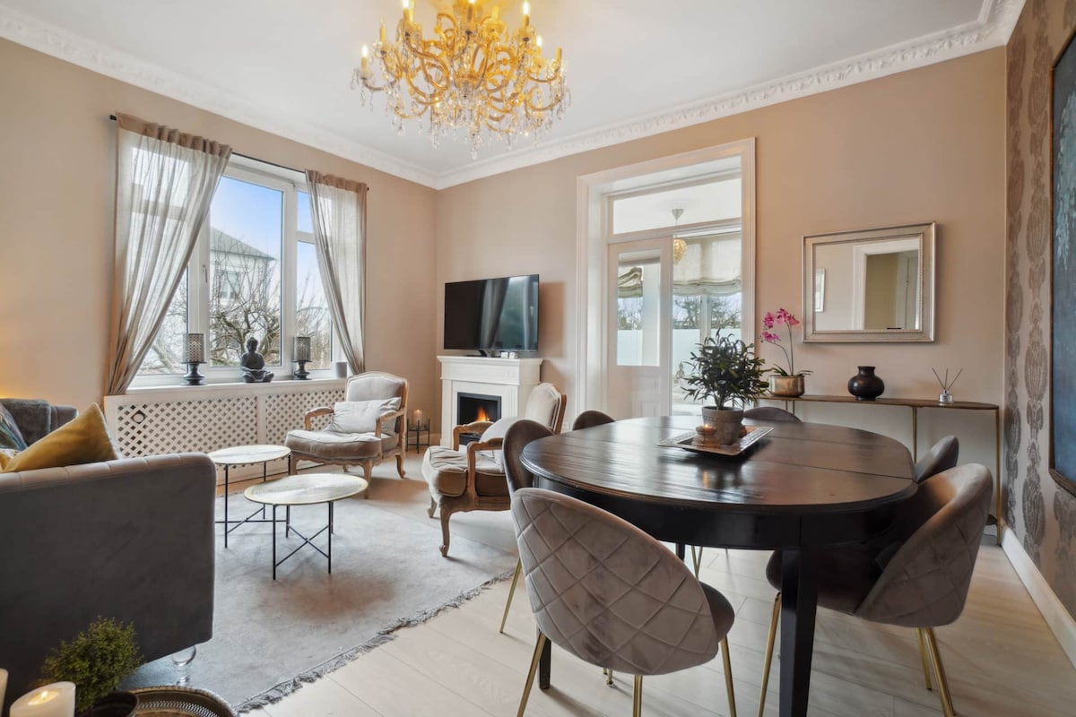 A well-arranged living area features elegant furnishings, including a round dining table surrounded by plush chairs. A chandelier hangs from the ceiling, and a cozy fireplace is nestled beneath a television. Natural light floods in through the window, enhancing the inviting ambiance.