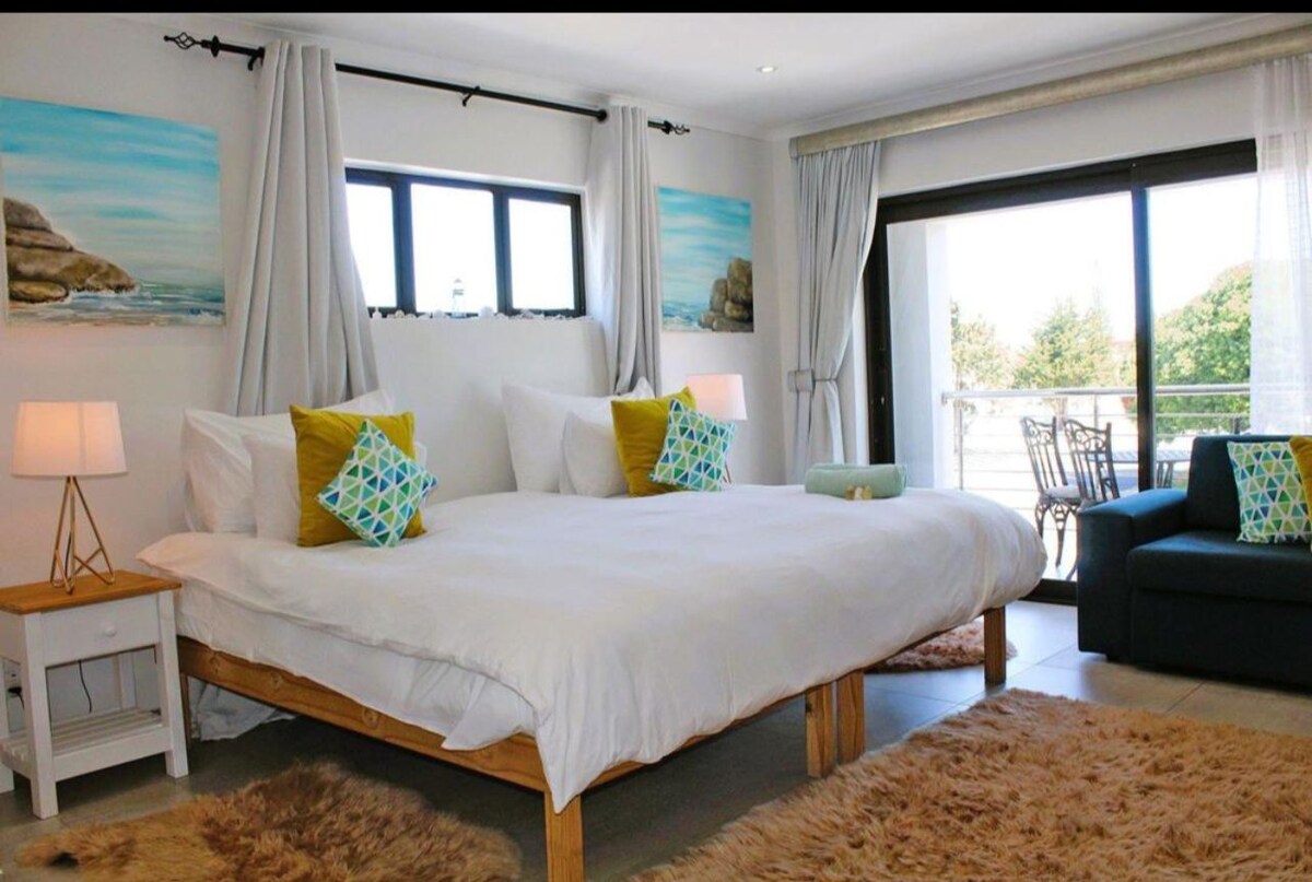 A bright and airy room features a spacious bed with fluffy white bedding and decorative pillows in refreshing colors. Two windows allow natural light to fill the space, complemented by light curtains. A cozy seating area is situated nearby, alongside bedside lamps and artwork portraying beach scenes.