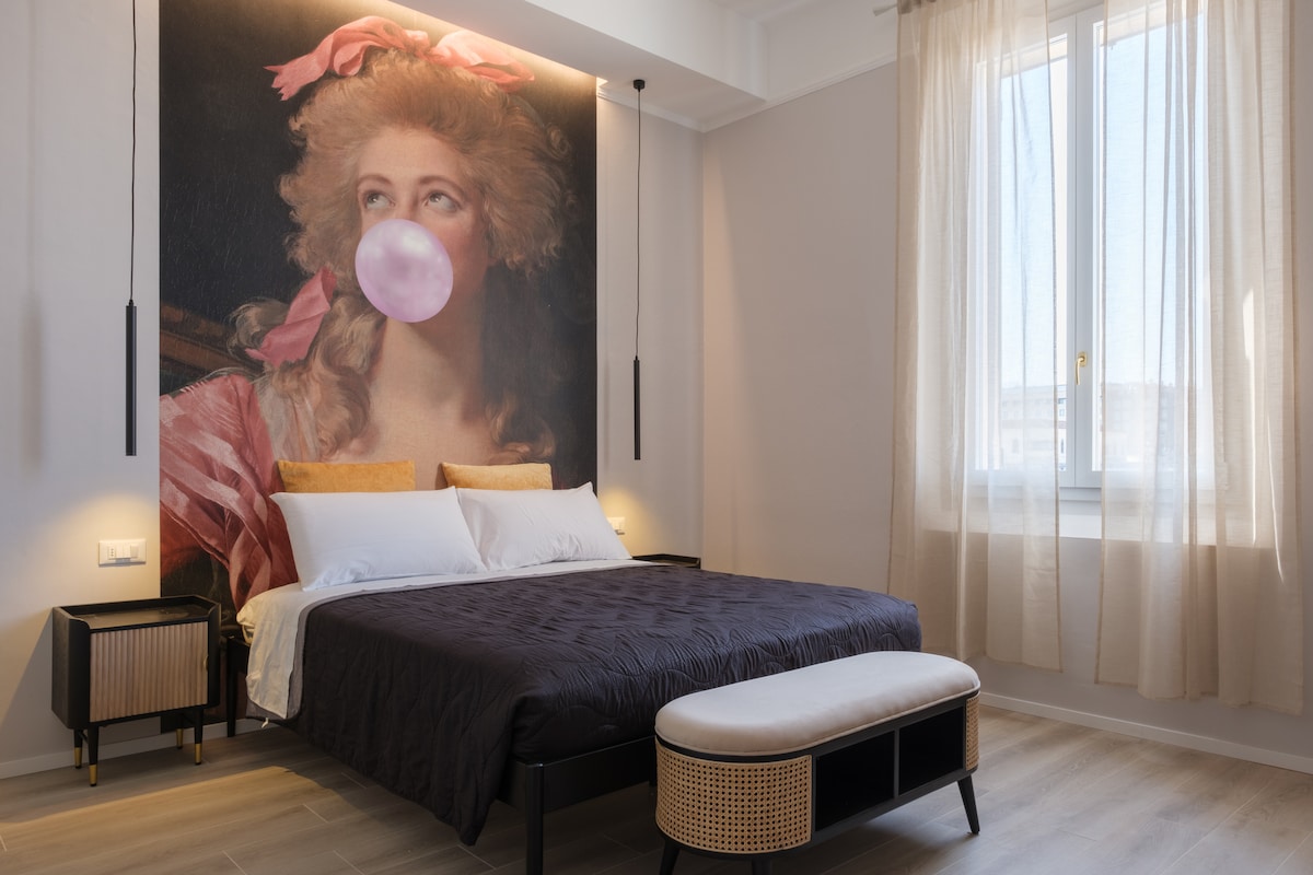 A tranquil bedroom is highlighted by a large artwork above the bed, featuring a woman's portrait. The spacious bed is dressed in smooth linens, and natural light filters through a large window adorned with sheer curtains. Warm tones from bedside lamps create a calming ambiance.