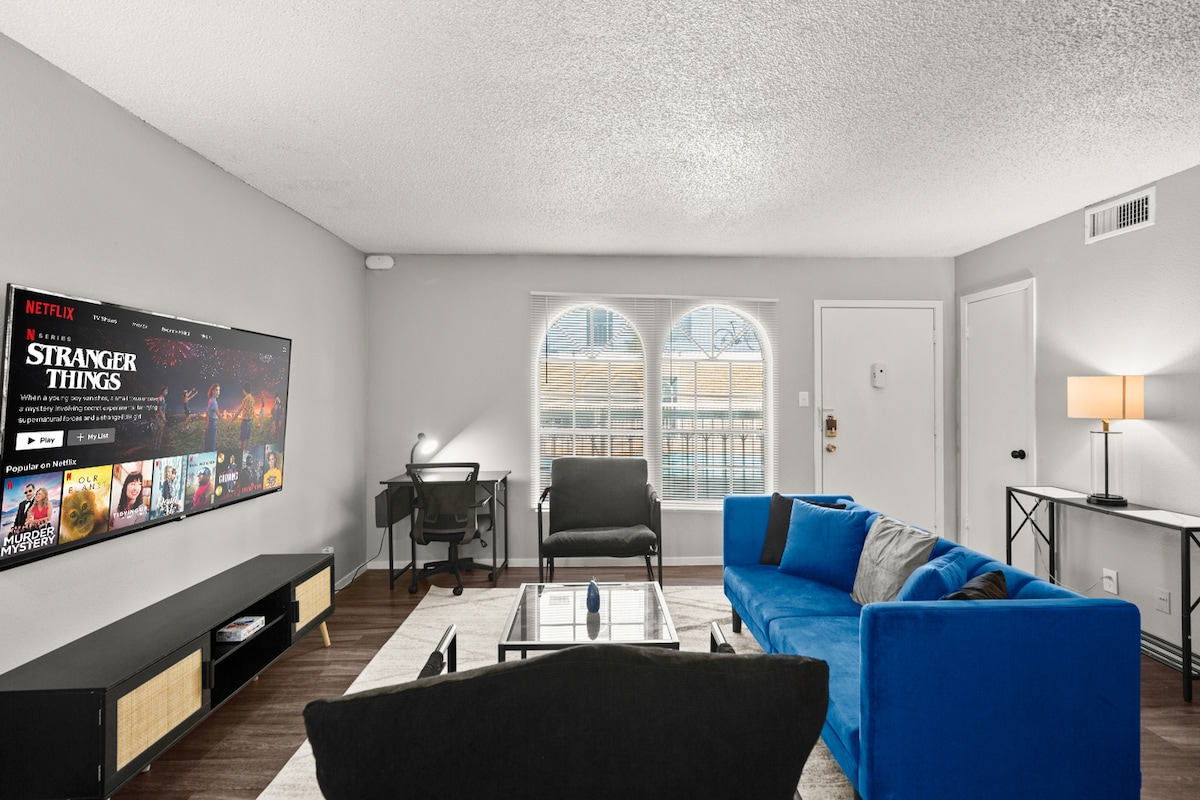 The living room features a large smart TV displaying popular streaming services, a chic coffee table, and a bright blue sectional sofa. Natural light floods the space through an arched window, complemented by a functional workspace equipped with a desk and chair.