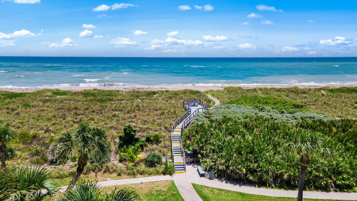 A scenic view of a pathway leading through lush greenery toward a sandy beach and the ocean. The vibrant blue waters meet a clear sky dotted with clouds, creating a serene coastal landscape.