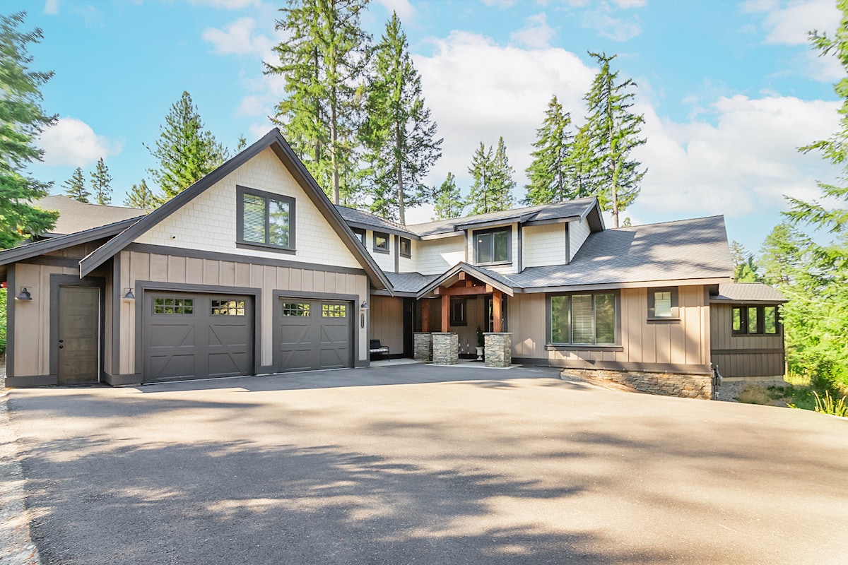 A spacious exterior of a modern retreat is presented, featuring a combination of wood and stone finishes. Large garage doors and a welcoming entryway are framed by tall trees, creating a serene and secluded atmosphere. Ample driveway space is available for parking.