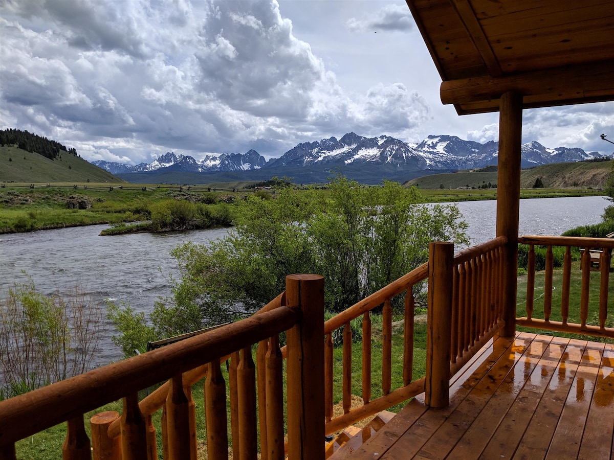 A covered deck with log railing overlooks lush greenery and flowing waters of the Salmon River. The backdrop features a panorama of the Sawtooth Mountains, partially shrouded in clouds, adding depth to the natural setting.