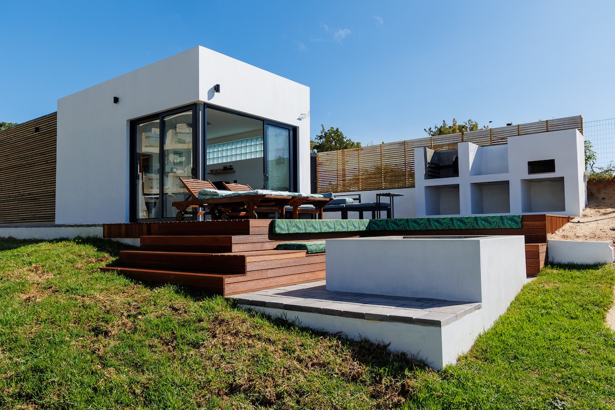 A modern structure is set against a clear blue sky, featuring large glass doors that open to a multi-level wooden deck. Outdoor seating is arranged around a stone fire pit, with a built-in barbecue area visible in the background.