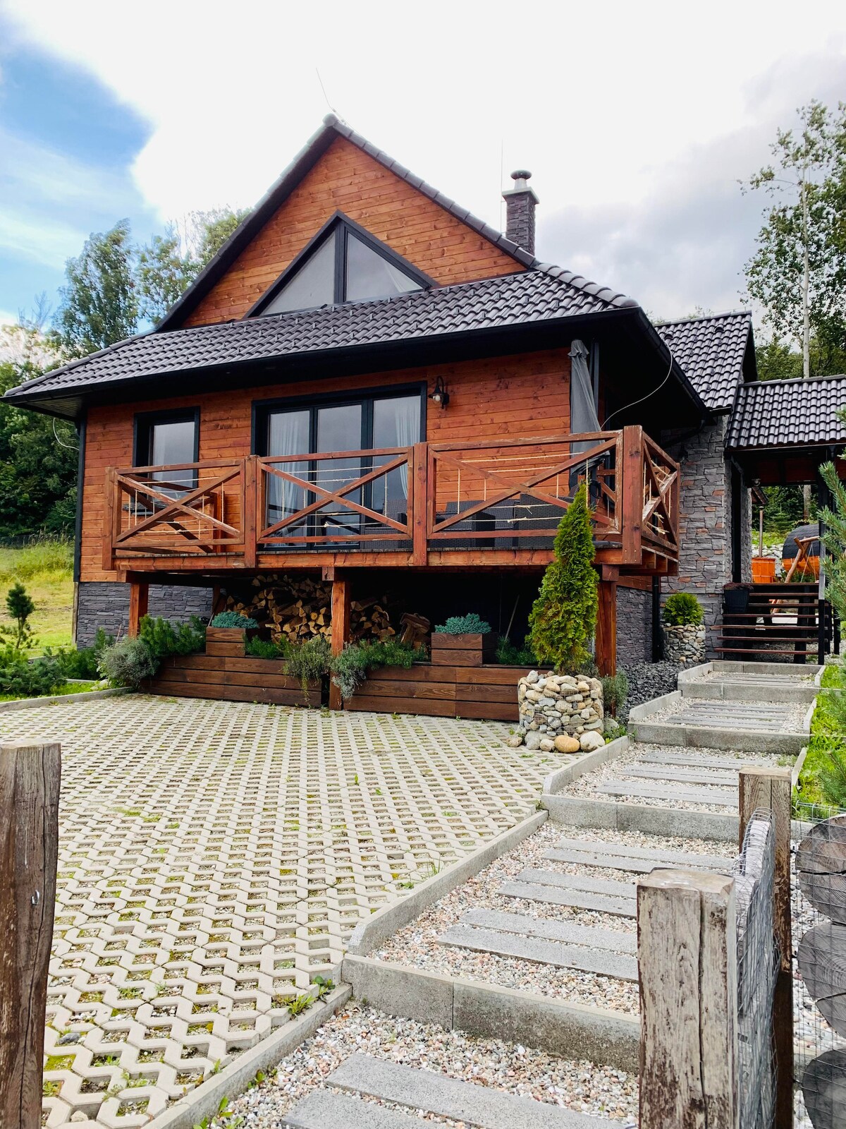 The exterior of the chalet features warm wooden panels, large windows, and a spacious balcony supported by wooden railings. Stone steps lead up to the entrance, surrounded by greenery and a well-maintained stone pathway.