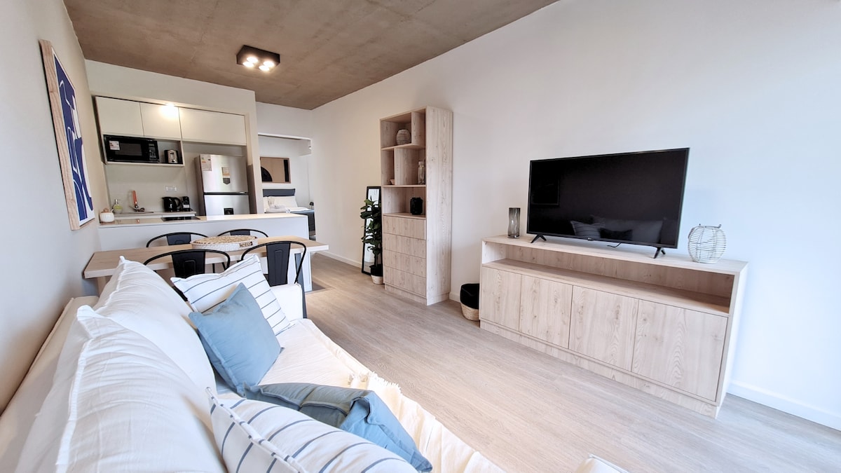 A modern living area features a comfortable sofa adorned with decorative pillows. A sleek television is placed on a wooden media console, with a small plant adding a touch of greenery. The open layout connects to a well-equipped kitchen in the background.