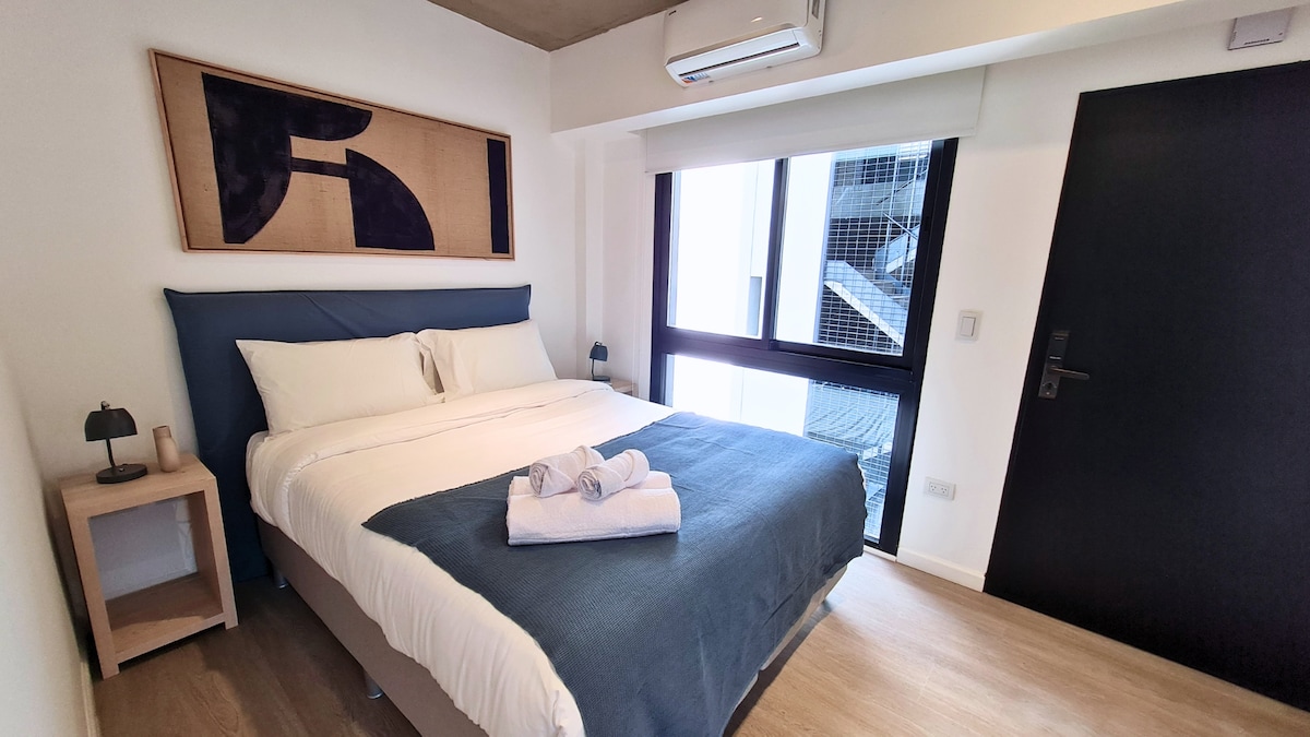 A well-lit bedroom features a spacious bed with a navy blue throw blanket and two neatly arranged towels. A contemporary artwork piece is displayed above the headboard. Large windows provide natural light, and a dark wooden door leads to the hallway.