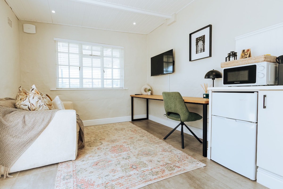 A bright living area features a cozy sofa adorned with patterned cushions. A small desk and chair are positioned against the wall, complemented by a flat-screen television. A microwave and mini fridge are included for added convenience, while a decorative rug adds a touch of warmth to the space.