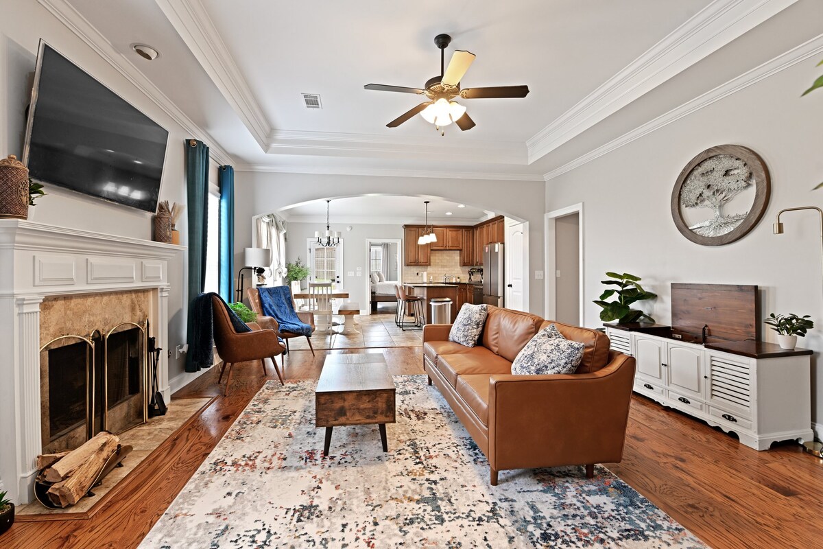 A spacious living area features a warm-toned leather sofa and a wooden coffee table atop a decorative rug. Natural light filters through large windows, illuminating the room. A television is mounted on the wall above a classic fireplace, while a cozy armchair is positioned nearby.