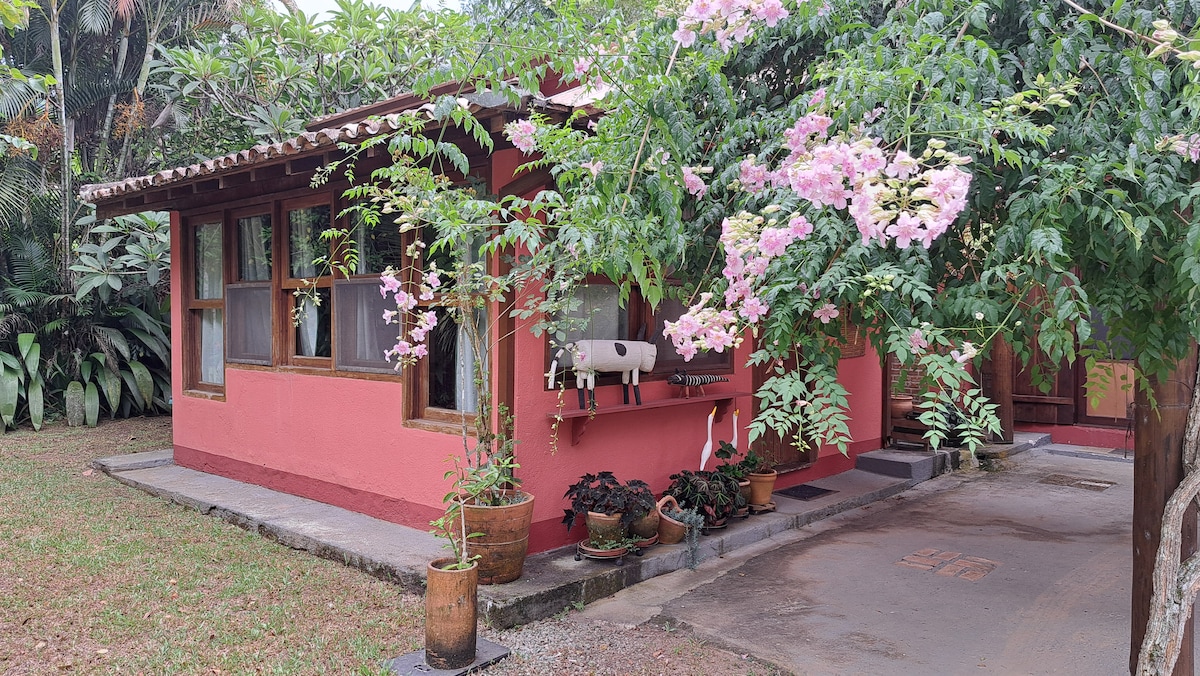 A charming red chalet is framed by green foliage and pink flowering plants, creating a serene outdoor setting. Large windows allow for natural light to enter, and potted plants are placed along the pathway, enhancing the inviting appearance of the exterior.