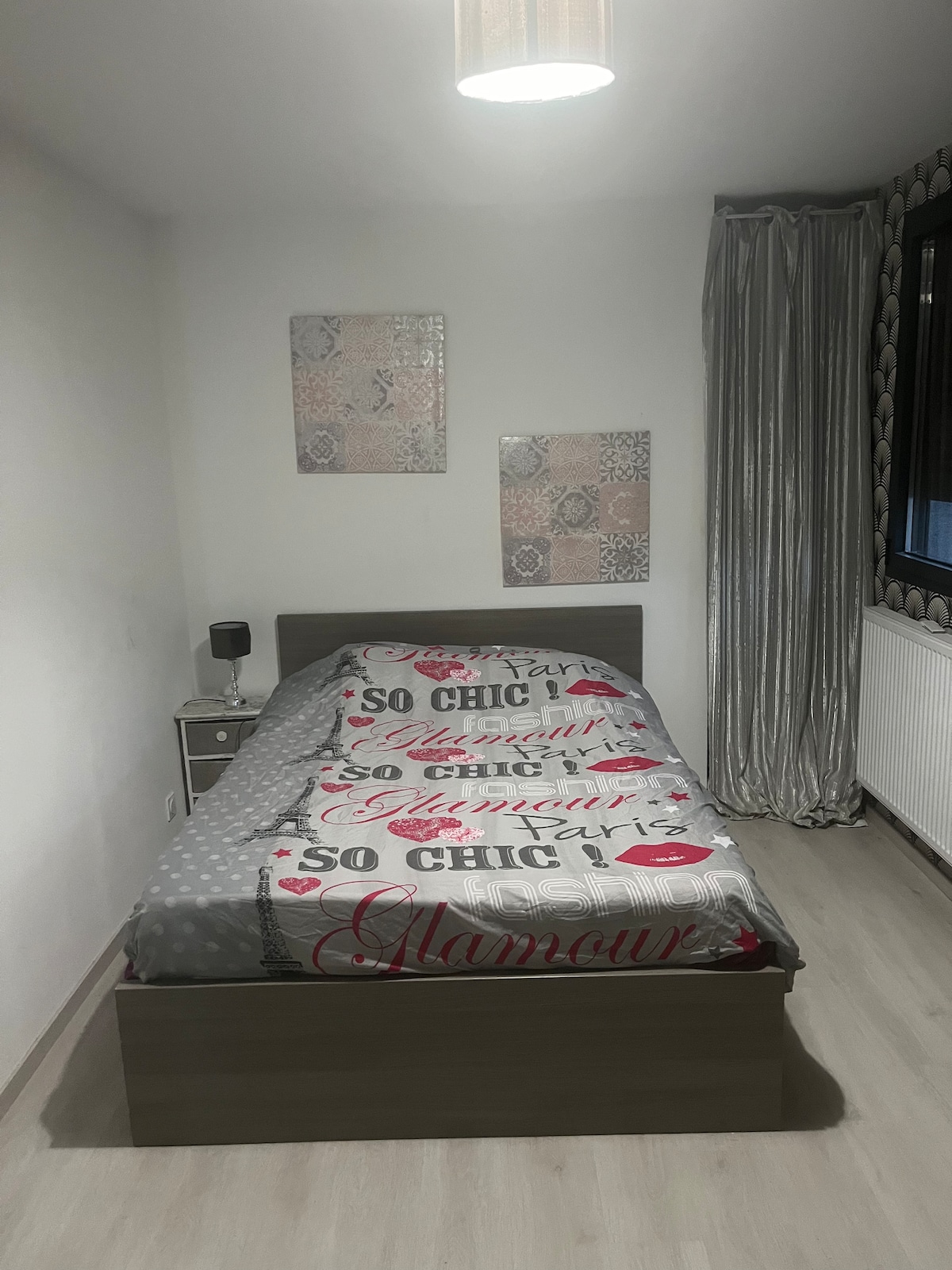 A cozy bedroom is presented, featuring a spacious bed covered with a decorative blanket. Two art pieces are displayed on the wall, adding visual interest. Soft curtains frame a large window, allowing natural light to filter into the minimalist space.