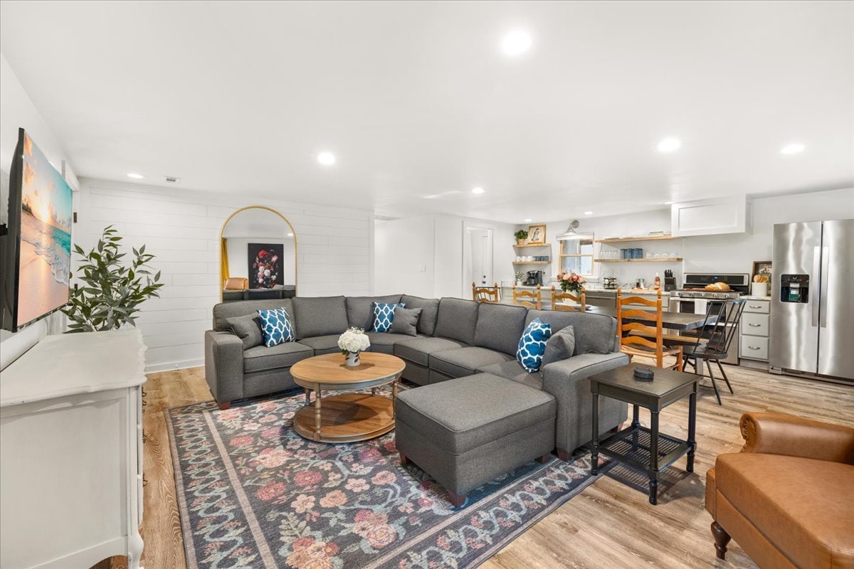 A spacious living area features a comfortable sectional sofa, a round coffee table, and a patterned area rug. A large TV is mounted on the wall, and the open kitchen is visible in the background, equipped with various appliances and dining options.
