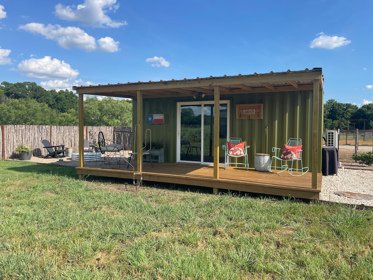 The exterior of The Bunkhouse is shown, featuring a covered wooden deck with seating options. Two colorful chairs are positioned at the front, alongside a small table. Large glass doors provide access to the interior, with a grassy yard and ranch landscape visible in the background.