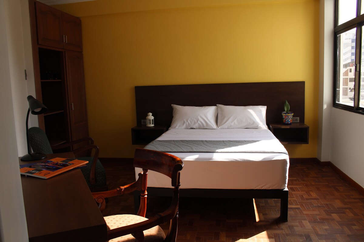 A well-lit bedroom features a spacious bed with crisp white linens against a bright yellow wall. A wooden bedside shelf houses a small plant, and a cozy chair is positioned near a desk. Large windows allow natural light to enhance the room's inviting atmosphere.