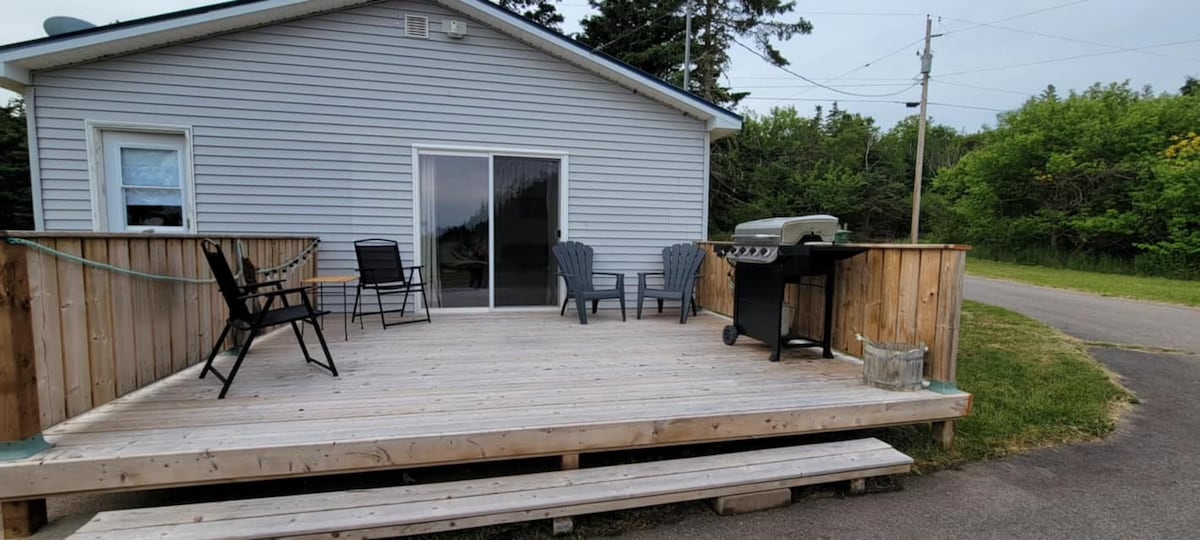 A spacious wooden deck features multiple seating options, including two comfortable chairs and a barbecue grill. Large glass doors open to the interior of the cottage, while the surrounding greenery adds a natural element.