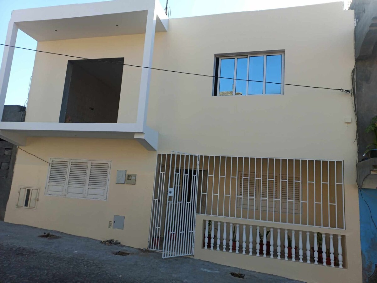 A two-story building is depicted, featuring a light-colored façade. Large windows adorn the upper level, while the lower level has a barred entrance and an adjacent balcony with white railings. The structure has a simple, modern design, set on a narrow street.
