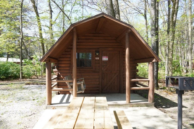 The rustic exterior of the cabin features weathered wooden walls and a sloped roof. A small porch with a swing offers a relaxing spot, while the entrance door is framed by natural surroundings, providing a welcoming view of the serene campground.