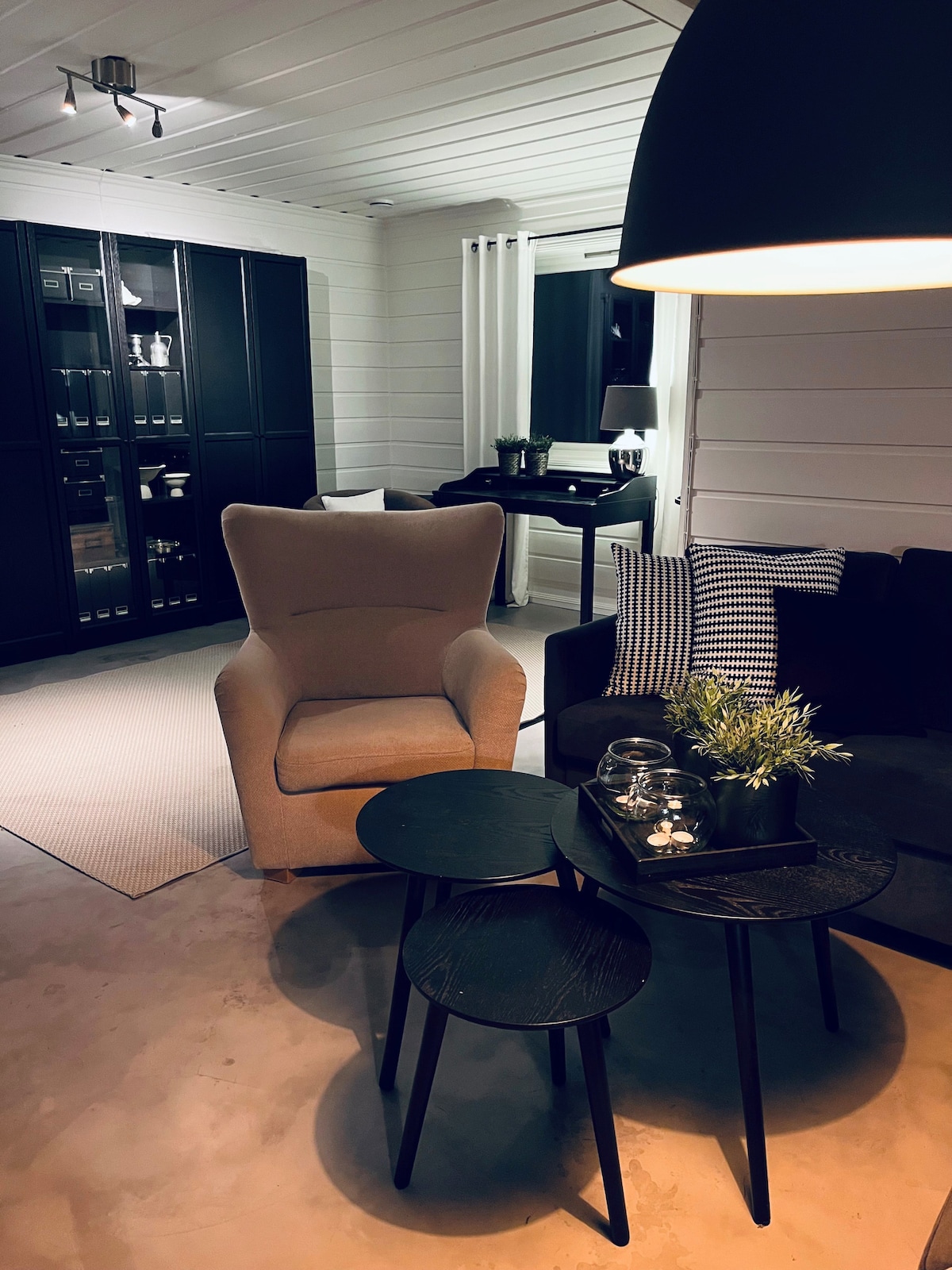 A cozy living area features a light-colored armchair alongside a dark sofa, creating an inviting seating arrangement. Two small, round tables are positioned in front, adorned with a potted plant and decorative items. The space is illuminated by a large pendant light, enhancing its warmth.