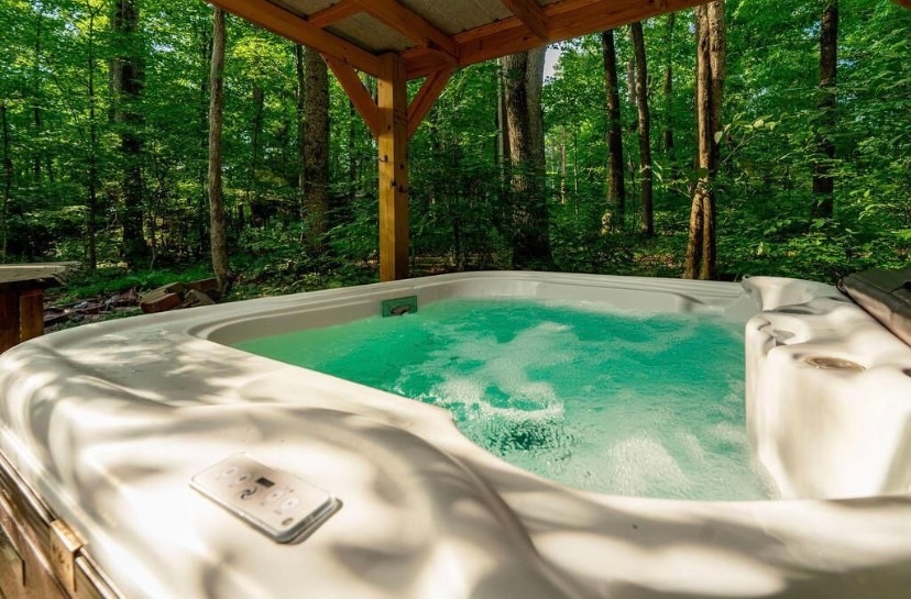 A hot tub is nestled under a wooden shelter, surrounded by lush greenery. Water bubbles gently within the tub, reflecting the dappled sunlight filtering through the forest canopy. The natural setting creates a serene atmosphere perfect for relaxation.