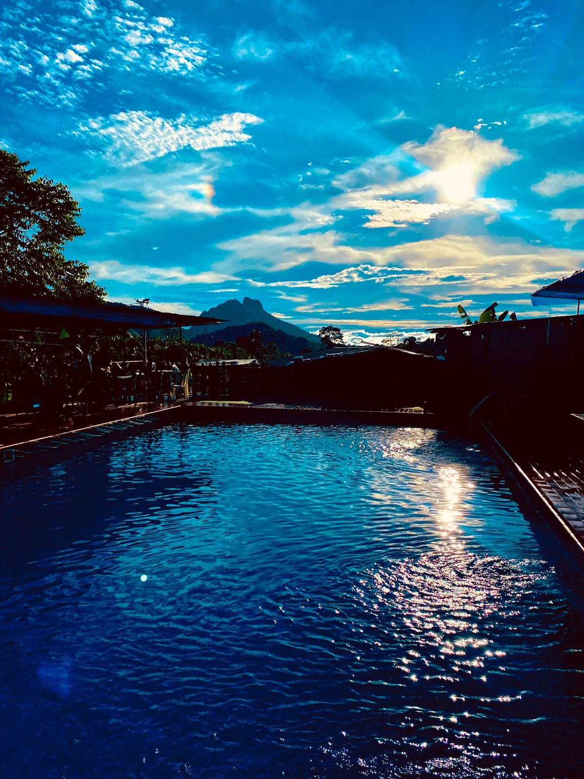A serene swimming pool reflects the vibrant sky, where shades of blue and orange merge during sunset. Lush greenery surrounds the area, enhancing the natural atmosphere. The mountains in the distance are silhouetted against the intricately clouded sky.