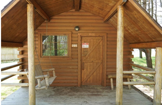 The cabin's rustic exterior features weathered wood logs and a welcoming porch. A wooden swing hangs at one side, inviting relaxation. A single window allows natural light, while the solid wood door has a simple design and a number visible on it.