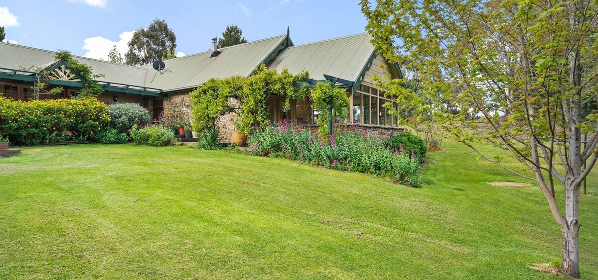 The exterior showcases a stone estate set amidst expansive lawns, with vibrant flower gardens and lush greenery surrounding the structure. A sunroom with large windows invites ample natural light. A few trees provide shade, creating a serene ambiance typical of the Australian bush.