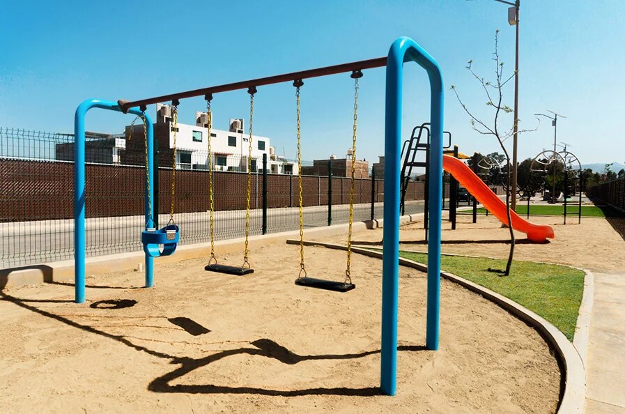 A playground area features a set of blue swings with black seats secured by yellow chains. An orange slide is positioned nearby, and a light tan sandy surface is visible, offering a play area for children. Fencing and green grass outline the space.