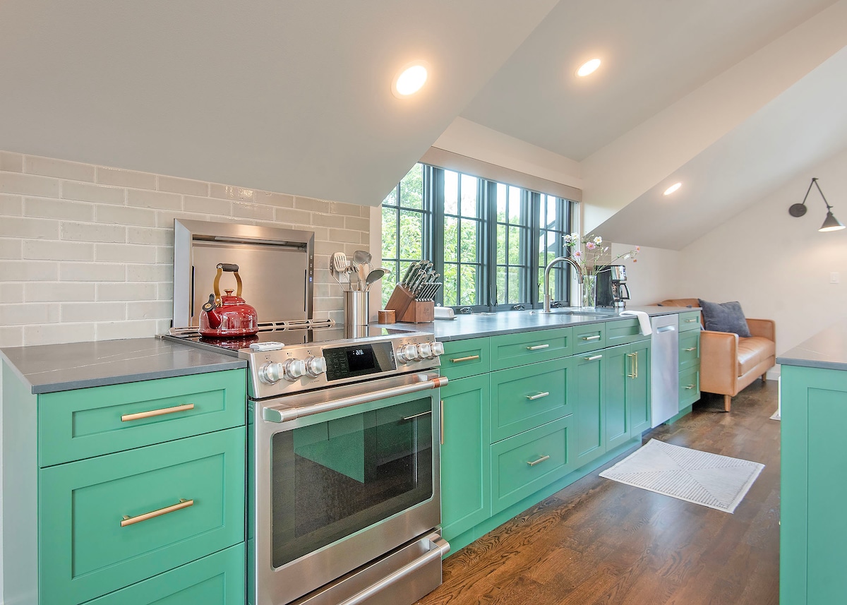 A modern kitchen features bright green cabinetry with gold hardware and sleek countertops. A stainless-steel stove is positioned beside a kitchen sink, with natural light streaming in through large windows. A cozy seating area is visible in the background, enhancing the inviting space.
