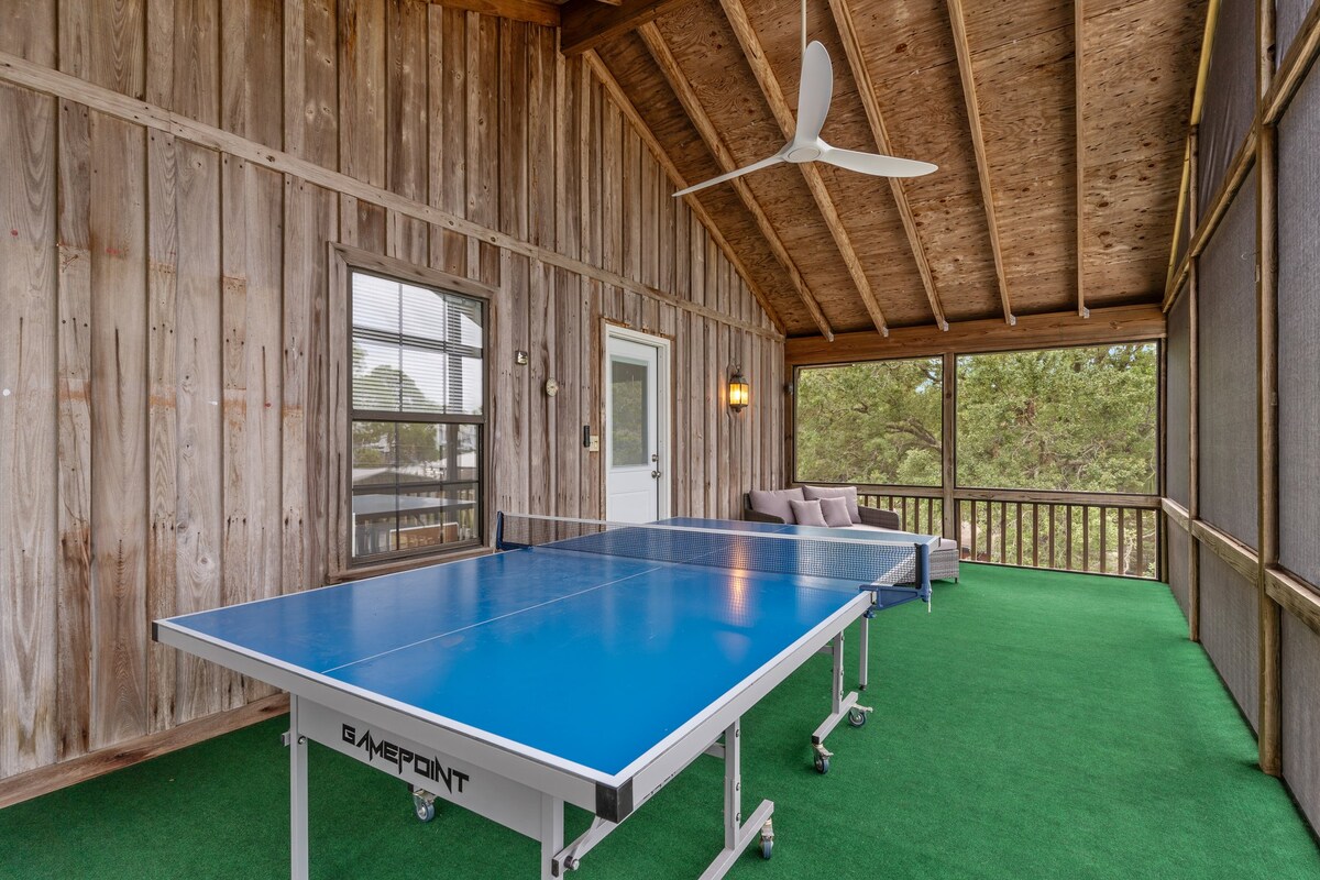 A screened porch features a blue ping pong table at its center, surrounded by wooden walls and a ceiling with exposed beams. A comfortable seating area is included, with a couch positioned against one side. Soft greenery covers the floor, enhancing the inviting space.