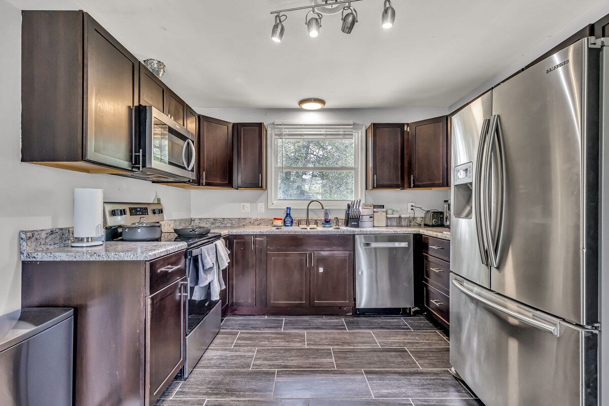 A modern kitchen features dark wood cabinetry and a stainless steel refrigerator. Countertops are finished in a speckled gray stone, complemented by a black stove and microwave. Natural light enters through a window, illuminating the space where various kitchen appliances and utensils are neatly arranged.