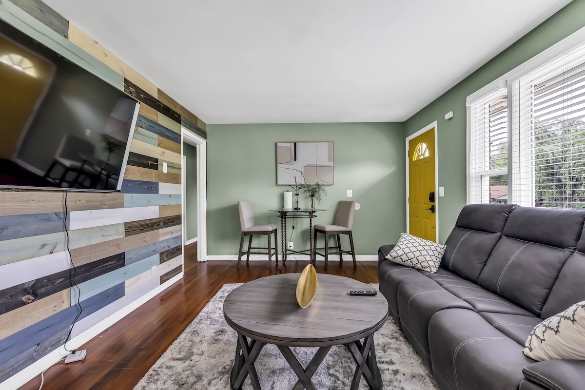 A comfortable living area features a modern sectional sofa and a round coffee table at the center. Two chairs are placed at a small dining table against a textured wood accent wall. Natural light enters through large windows, complemented by vibrant green walls.