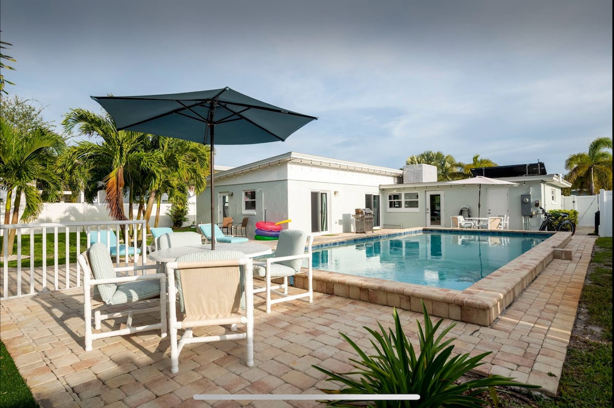 An outdoor space is showcased, featuring a pool surrounded by a stone patio. A shaded dining area is furnished with a light-colored table and chairs. Lush greenery and palm trees provide a tropical backdrop, contributing to a relaxing atmosphere.