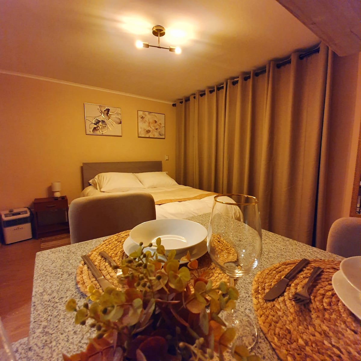 A cozy dining area is featured in the foreground, with a neatly set table adorned with plates and transparent glasses. In the background, a spacious bed is partially visible, surrounded by soft curtains, complemented by minimal wall art above the bed.