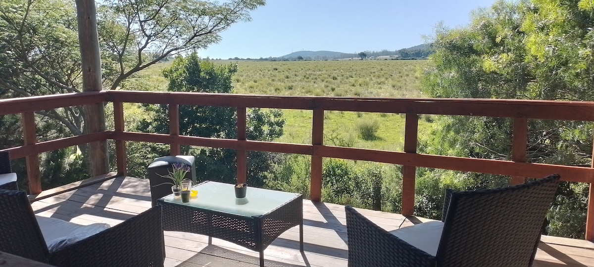 A wooden deck offers a wide view of the surrounding fields and mountains. Two wicker chairs sit beside a glass coffee table, complemented by a small plant and a candle. The area is bathed in natural light, surrounded by greenery.
