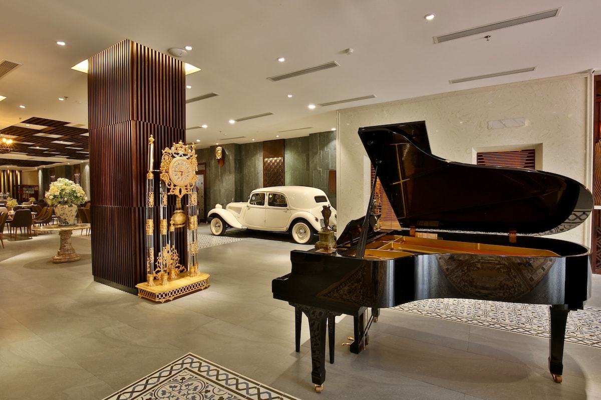 An elegant lobby space features a grand piano with polished black finishes, positioned near a vintage car. Decorative elements include a prominent gold clock and tasteful floral arrangements. The area is illuminated with warm lighting, enhancing the sophisticated ambiance.