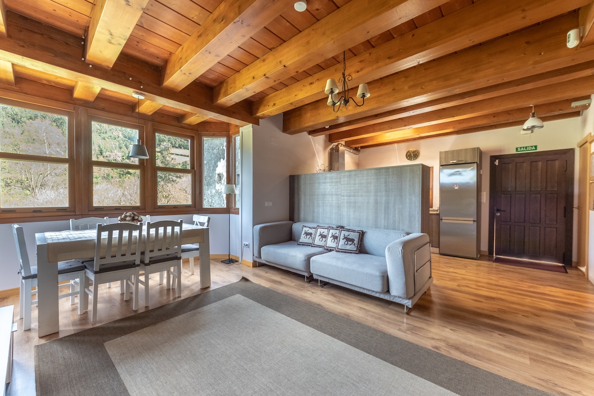 An open-plan living area features wooden beams overhead and large windows allowing natural light. A comfortable gray sofa is positioned near the dining table, which accommodates six. Light wooden flooring extends throughout, contributing to a relaxed atmosphere. A modern kitchen area can be seen in the background.