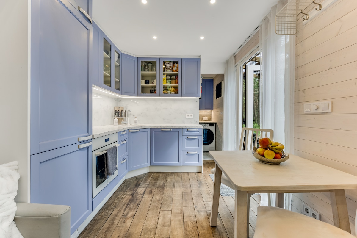 A modern kitchen is shown, featuring light blue cabinetry and white marble countertops. A small dining table is set beside a window, with a bowl of fresh fruit placed on top. Natural wood flooring adds warmth to the space.