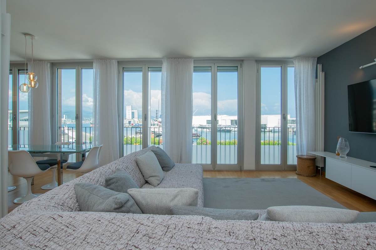 A spacious living area is highlighted by large glass doors that provide an unobstructed view of the harbor. Natural light floods the room, which features a comfortable sectional sofa and a dining area surrounded by modern chairs. Sheer curtains gently frame the windows.