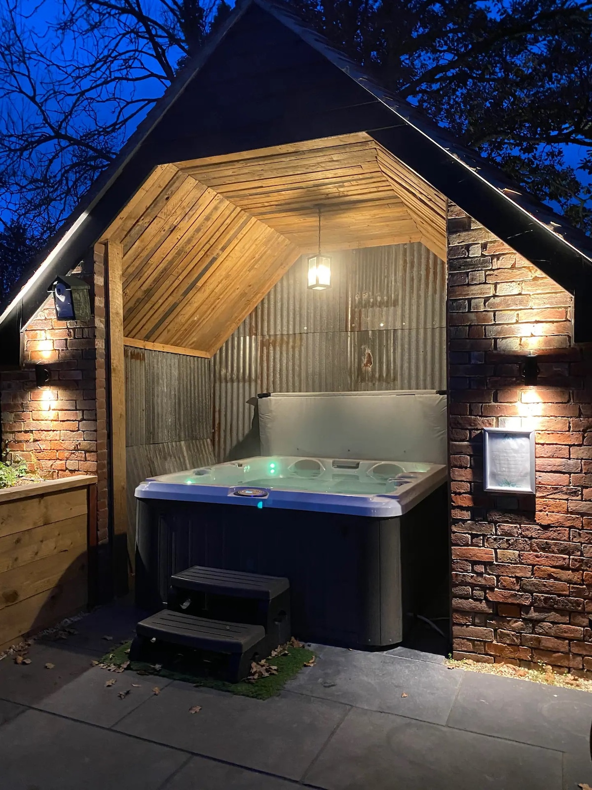 A private hot tub is situated within a cozy wooden structure, featuring rustic brick walls and a gabled roof. Soft lighting enhances the ambiance, creating a serene atmosphere ideal for relaxation. A small step leads up to the hot tub, framed by natural elements.