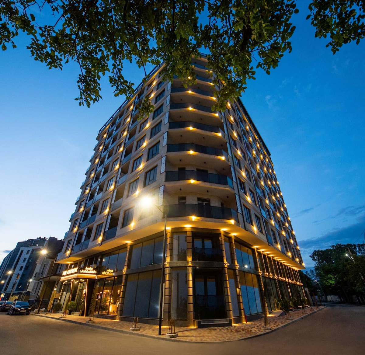 The modern Gori Palace building is illuminated at dusk, showcasing its sleek architecture with prominent balconies and large windows. Soft lighting highlights its facade while trees frame the image, contributing to the inviting streetscape.