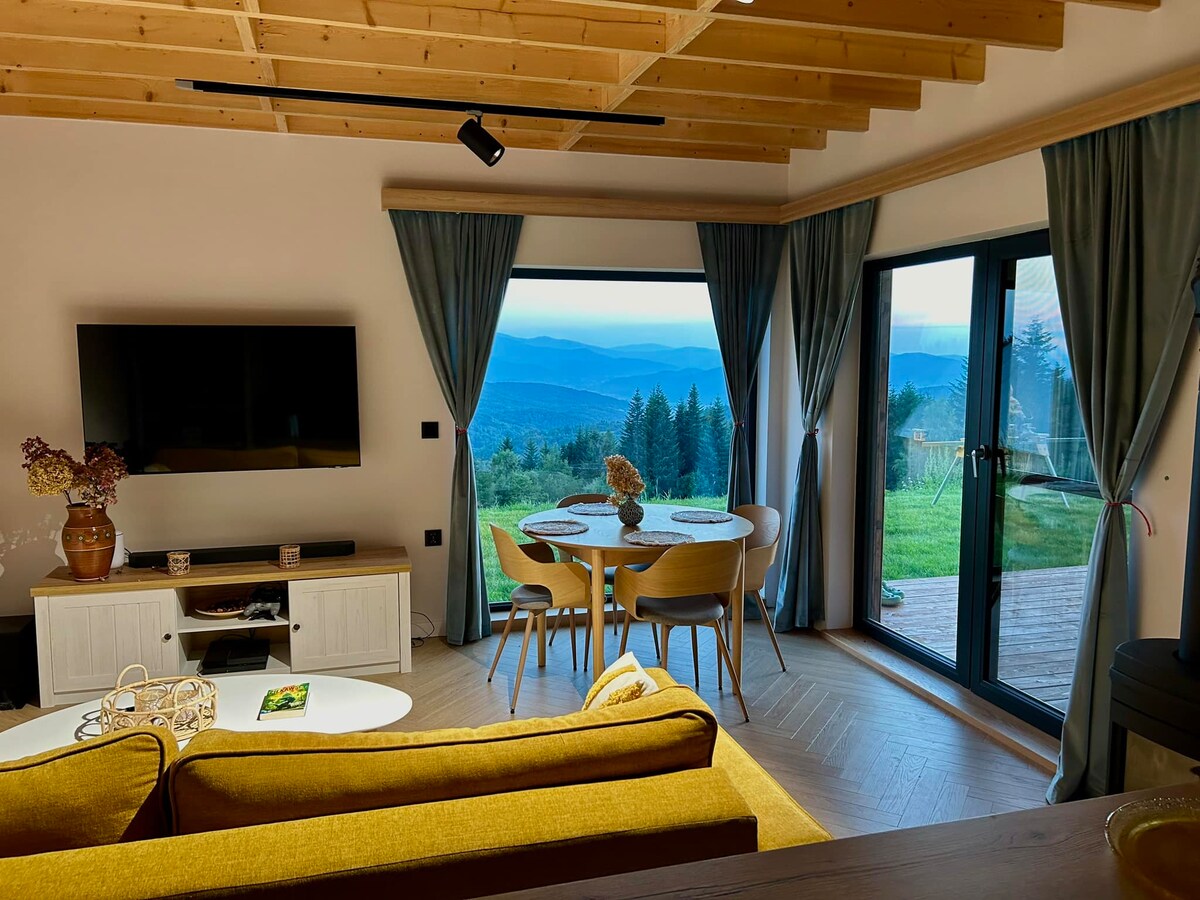 The living room features large glass doors that open to stunning mountain views. A comfortable yellow sofa invites relaxation, while a stylish TV unit displays a flat-screen television. A round dining table with chairs is positioned near the window, creating a bright dining area with natural light.