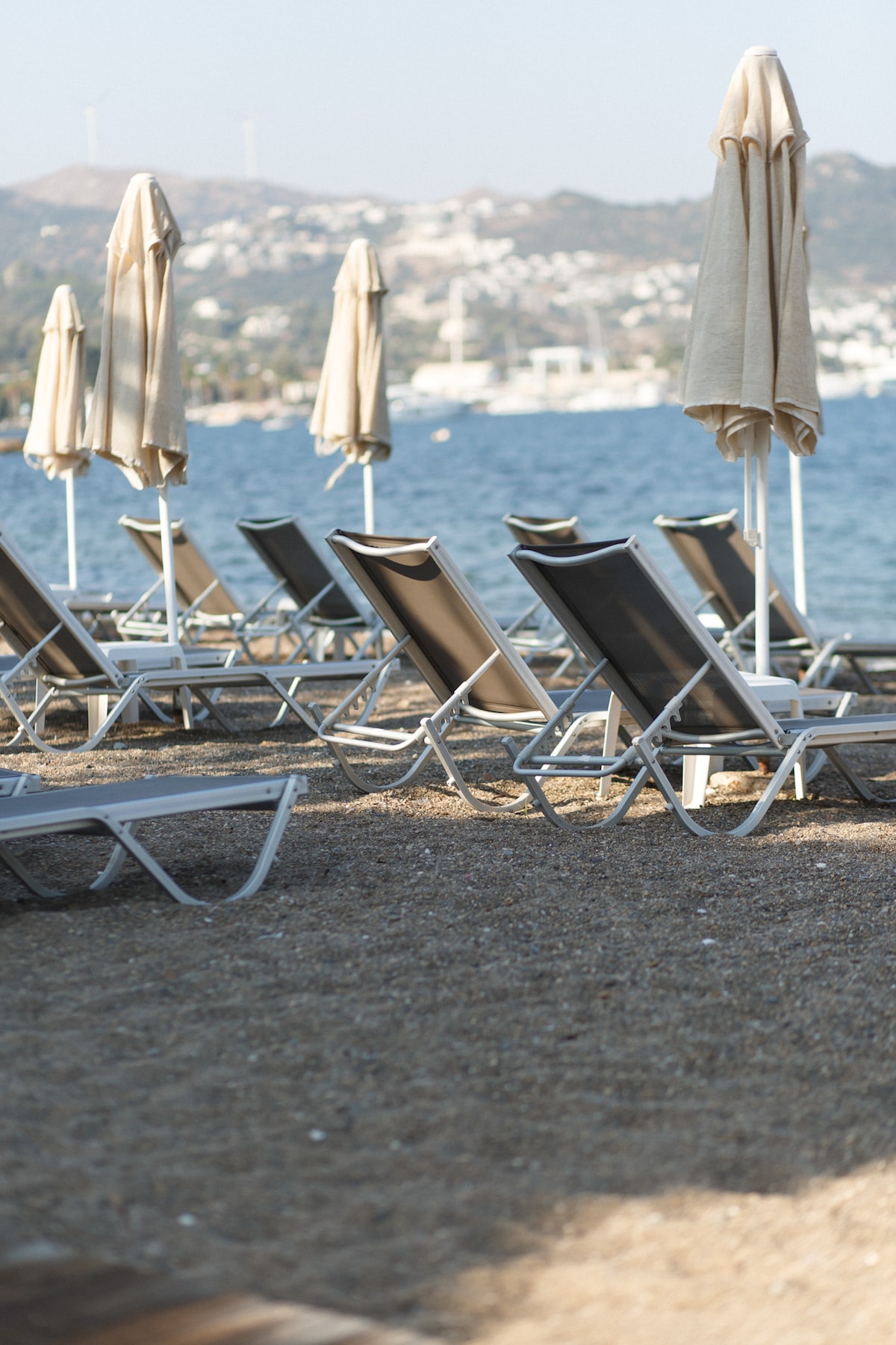 A row of lounge chairs is positioned on a sandy beach, accompanied by tall umbrellas providing shade. The tranquil sea is visible in the background, with distant hills dotting the landscape.