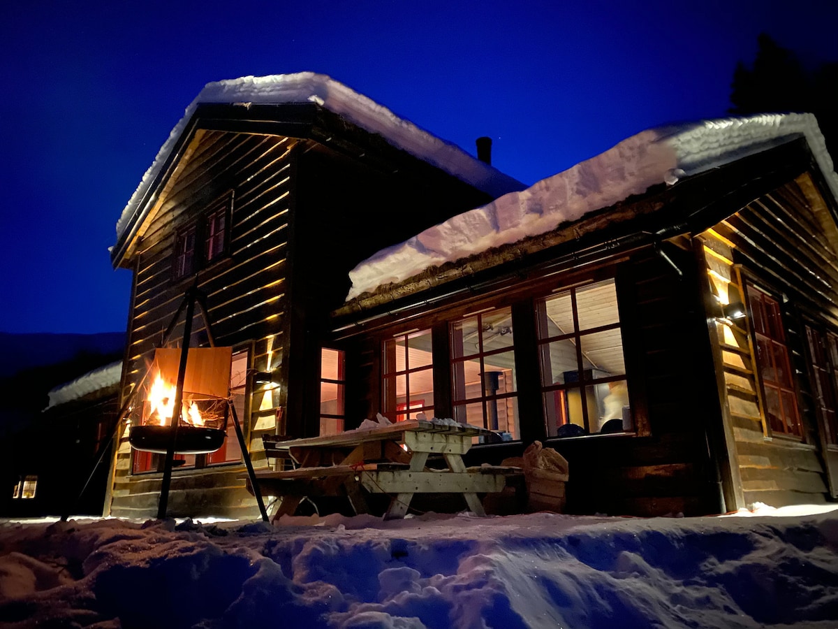 A wooden cabin is illuminated at night, with snow covering the roof and surroundings. Warm light spills from large windows, creating a cozy atmosphere. An outdoor seating area features a picnic table, and a fire pit is positioned nearby, enhancing the inviting setting.