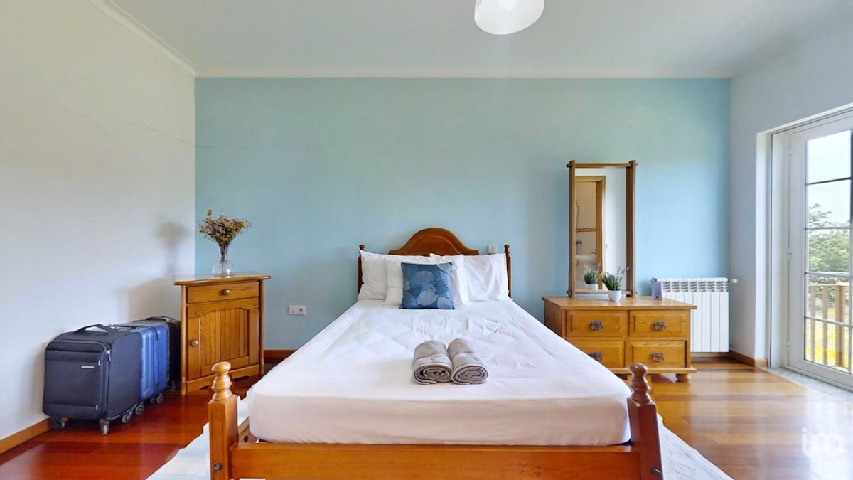 A serene bedroom features a spacious bed with white linens and two rolled towels placed on top. A wooden nightstand and dresser are positioned beside the bed. Luggage is nearby, and natural light enters through a large window, complemented by light blue walls.
